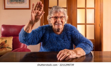 Happy Old Woman With A Smile On Her Face Fallen Hand In Greeting. Caucasian 90s Grandma In Living Room At Home. Grandmother Hello And Welcoming Guests Concept.