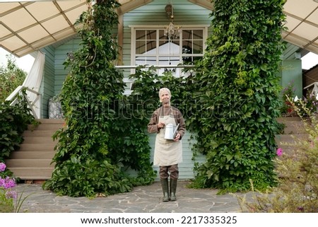 Happy old woman in workwear standing in front of camera against cottage or summer house overgrown with green grape leaves
