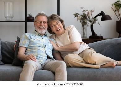 Happy Older 60s Couple Relaxing In Living Room Home Portrait. Senior Husband And Wife Resting, Hugging On Couch, Looking At Camera, Laughing, Smiling, Talking On Video Call. Screen View