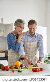 Happy older 60s vegan couple wearing aprons preparing healthy diet meal salad at home. Smiling mature senior family embracing, cooking together, cutting fresh organic vegetables together in kitchen.