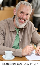 Happy older adult business man professor or student writing in notebook, smiling middle aged gray-haired bearded author or writer taking notes looking at camera sitting at table. Vertical portrait.