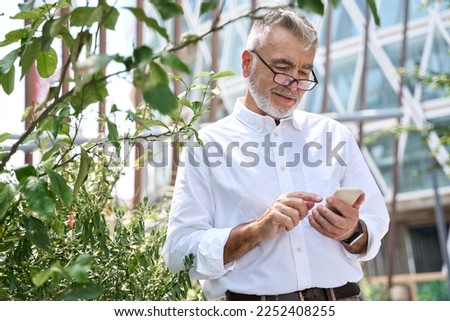 Happy older adult professional business man, smiling senior 60 years old businessman holding smartphone, using mobile digital tech apps checking cell phone standing in city park outdoors.