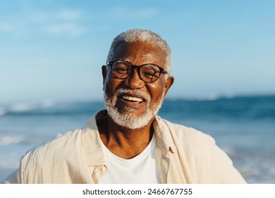 A happy older African American man with glasses and a white beard enjoys the ocean breeze on a sunny day at the beach, portraying happiness and contentment. - Powered by Shutterstock