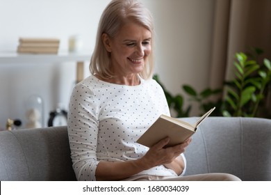 Happy older blonde woman sitting on comfortable sofa in living room, reading interesting book. Smiling elder retired grandmother enjoying leisure weekend time with favorite novel alone at home.