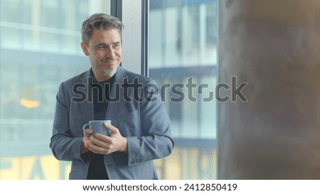 Happy older businessman taking break in office drinking coffee standing in window and daydreaming, thinking.