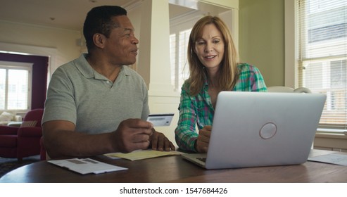 Happy Older Couple Booking Trip Or Making Purchase Online With Credit Card Using Laptop Computer. African American And Caucasian Husband And Wife Paying Off Outstanding Debt