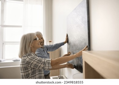 Happy older couple hanging picture on wall at home, enjoying moving into new home after renovation, decorating apartment together, smiling, laughing, discussing abstract artwork - Powered by Shutterstock