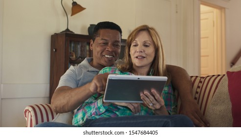 Happy Older Couple On The Couch Using Tablet Computer Together To Watch Funny Video. African American And Caucasian Husband And Wife Playing Games Or Streaming With Mobile Device