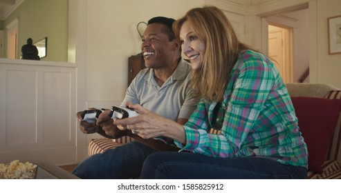 Happy Older Couple Playing Video Games Together On Living Room Couch. African American And Caucasian Husband And Wife Using Console Controllers To Play Videogame