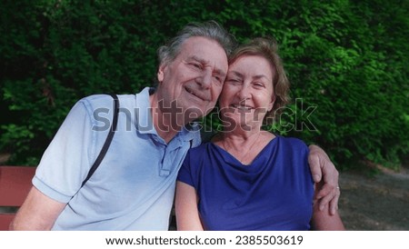 Happy older couple sitting at park bench during sunny summer day. Senior husband putting arm around elderly wife posing together, close-up faces, cheek to cheek