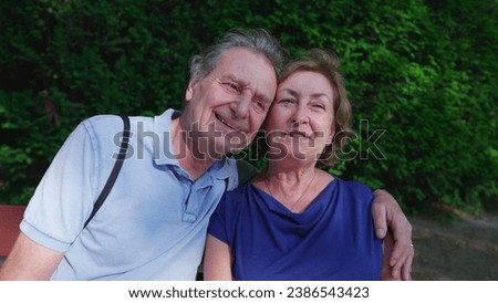 Happy older couple sitting at park bench during sunny summer day. Senior husband putting arm around elderly wife posing together, close-up faces, cheek to cheek
