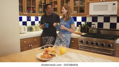 Happy Older Couple Talking And Drinking Morning Coffee In Their Kitchen. Happily Married Black And White Husband And Wife Chatting About Their Day Wearing Pajamas