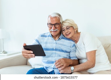 Happy Older Couple Using Computer Tablet Together At Home, Excited Mature Man And Woman Looking At Mobile Device Screen, Shopping Or Chatting Online, Sitting On Cozy Couch In Living Room