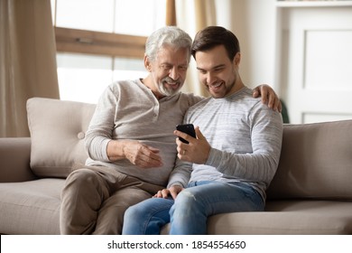 Happy Older Father And Adult Son Hugging, Using Phone At Home Together, Smiling Mature Grandfather And Grandson Looking At Screen, Browsing Apps, Young Man Teaching Senior Dad To Use Smartphone