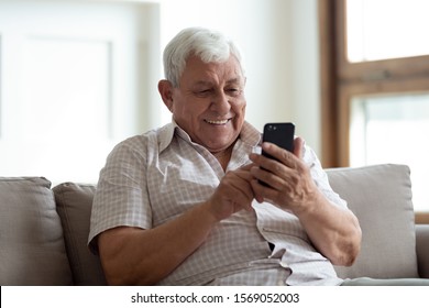 Happy older man sitting on comfortable sofa at home, using smartphone. Pleasant 80s grandfather reading sms with good news, typing message for grown up kids, shopping in mobile app, watching photos.