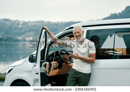 Happy older man standing near rv camper van on vacation using mobile phone. Smiling mature active traveler holding smartphone enjoying free roaming or internet in camping tourism nature park.