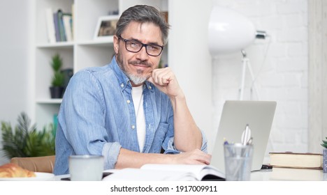 Happy older man working online with laptop computer at home sitting at desk. Home office, browsing internet, study room. Portrait of mature age, middle age, mid adult man in 50s.