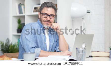 Happy older man working online with laptop computer at home sitting at desk. Home office, browsing internet, study room. Portrait of mature age, middle age, mid adult man in 50s.