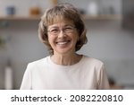 Happy older mature 60s woman indoor head shot portrait. Cheerful elder lady wearing eye glasses, casual, looking at camera with toothy smile, posing at home, laughing. Video call screen