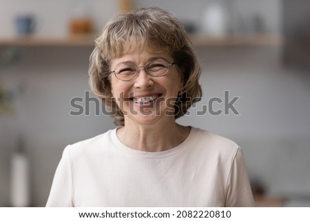 Happy older mature 60s woman indoor head shot portrait. Cheerful elder lady wearing eye glasses, casual, looking at camera with toothy smile, posing at home, laughing. Video call screen