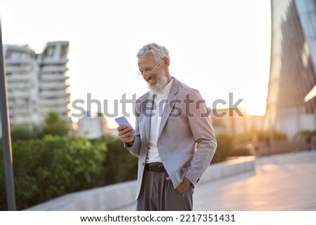 Happy older mature adult professional business man, smiling senior old businessman wearing suit holding smartphone using mobile cell phone standing outdoor in big city downtown among office buildings.