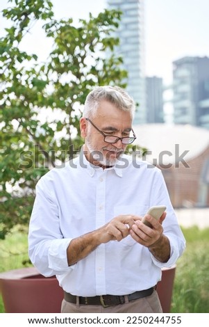 Happy older mature adult professional business man, smiling senior old bearded businessman executive holding smartphone, using mobile digital technology cell phone standing in city park, vertical.