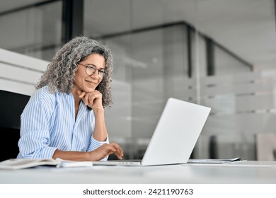 Happy older mature business woman entrepreneur in office using laptop at work, smiling professional senior middle aged female executive wearing glasses working on computer at office workplace desk.