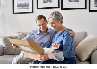 Happy Older Mature Couple Customers Unpacking Parcel Sitting At Home On Couch. Senior Middle Aged Grandparents Opening Online Store Order Receiving Gift In Postal Delivery Shipping Box.