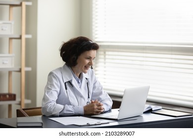 Happy older mature female doctor therapist physician in headset with mic looking at laptop screen, consulting patient remotely by distant web camera video call meeting in clinic, telemedicine concept.