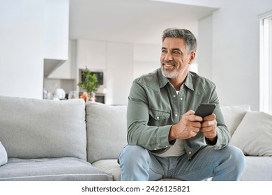 Happy older mature middle aged man holding cell phone using smartphone sitting at home on sofa, relaxing on couch in living room, looking away at copy space advertising mobile offers and apps. - Powered by Shutterstock