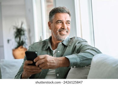 Happy older mature middle aged man holding cell mobile phone using smartphone sitting at home on sofa, relaxing, scrolling social media, checking financial apps, buying online, looking away at window. - Powered by Shutterstock