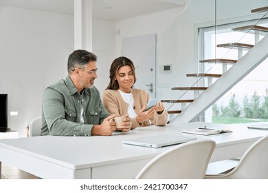 Happy older mature senior family couple, middle aged man and woman looking at cell phone using smartphone mobile technology device together and talking sitting at home table doing online shopping. - Powered by Shutterstock