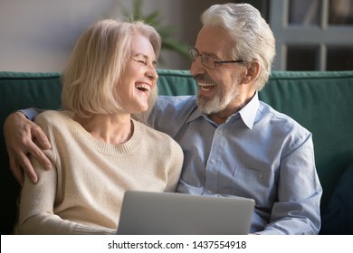 Happy Older Mature Spouses Couple Laughing Embracing Using Laptop At Home, Positive Cheerful Senior Middle Aged Family Husband And Wife Bonding Having Fun With Computer Sitting On Couch Together