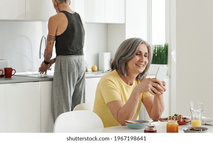 Happy Older Mature Woman Wife Using Mobile Apps On Cell Phone Ordering Food Delivery Shopping In Web Store Holding Smartphone Sitting At Kitchen Table While Senior Husband Washing Dishes At Home.