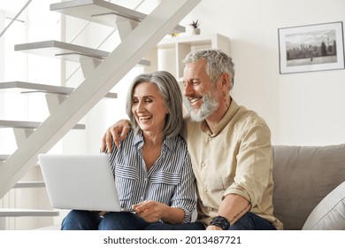Happy Older Mid Age Family Couple Using Laptop Sit On Couch. Smiling Senior Adult Mature Man And Woman Looking At Computer Doing Ecommerce Online Shopping, Watching Tv, Having Virtual Chat At Home.