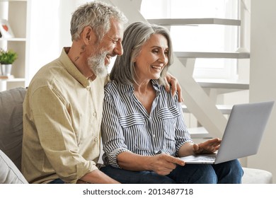 Happy Older Middle Age Family Couple Using Laptop Sit On Couch. Smiling Senior Adult Mature Man And Woman Looking At Computer Doing Ecommerce Online Shopping, Watching Tv, Having Virtual Chat At Home.
