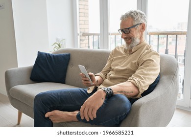 Happy older middle aged man using apps on phone relaxing sitting on couch at home. Smiling senior hipster with tattoos wearing glasses holding cellphone device ordering delivery online or texting.