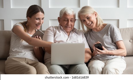 Happy Older Parents With Adult Daughter Using Electronic Devices, Phones, Looking At Laptop Screen, Smiling Grandparents And Granddaughter Watching Funny Video Or Reading News In Social Network