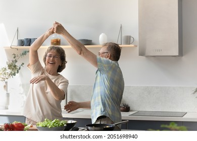 Happy older retired man twisting elderly wife, dancing to music in modern kitchen, taking break in preparing healthy food. Joyful laughing middle aged family couple having fun, enjoying hobby activity