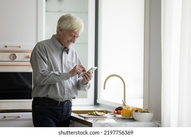 Happy Older Retired Man Using Mobile Phone Over Kitchen Counter, Chatting Online At Table With Fresh Vegetables For Salad, Organic Food, Natural Ingredients, Browsing Recipe On Internet