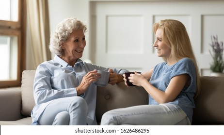 Happy Older Retired Woman Mother Chatting With Pleasant Smiling Grown Up Daughter, Sitting Together On Cozy Sofa, Drinking Tea Coffee. Loving Two Generations Family Enjoying Free Leisure Time At Home.