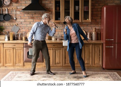 Happy Older Senior Loving Family Couple Having Fun In Kitchen, Singing Favorite Karaoke Songs In Utensils. Positive Joyful Middle Aged Spouse Enjoying Funny Domestic Weekend Activity Together Indoors.