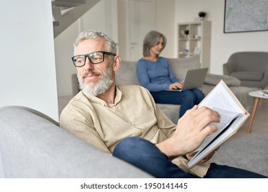 Happy Older Senior Man Husband Wearing Glasses Reading Book Relaxing Sitting On Couch At Home With Mature Wife Using Laptop. Middle Aged Family Couple Enjoying Casual Daily Activities