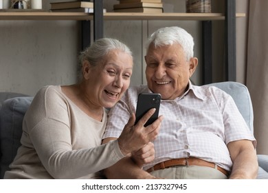 Happy older wife and husband laughing, have fun using modern smartphone, rest sit on sofa at home. Smiling senior couple read good news feel excited, get great offer, receive notice looking overjoyed