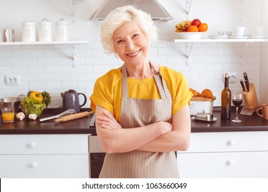 Happy older woman in apron preparing breakfast, crossed hands