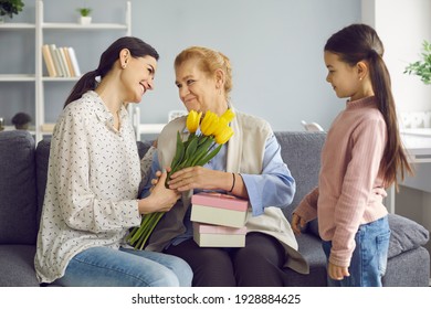 Happy Older Woman Receives Gifts In Honor Of Mother's Day Or International Women's Day. Loving Young Daughter And Little Granddaughter Give Old Mother And Grandmother Flowers And Gifts.