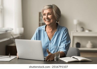 Happy older woman sits at desk with laptop, glass of water and open notebook nearby, feels satisfied while working, managing tasks in positive and productive atmosphere. Lifestyle, modern technology - Powered by Shutterstock