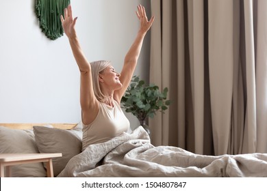 Happy older woman sitting in comfortable bed, stretching hands after awakening in bedroom, happy satisfied older female doing easy exercise in the morning, greeting new day, sunrise - Powered by Shutterstock