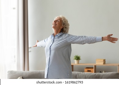 Happy older woman with toothy healthy smile standing with arms outstretched in living room at home, thankful grateful mature female feeling satisfied, doing easy exercises, stretching, enjoying life