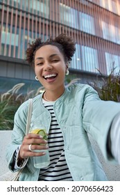 Happy optimistic girl with two hair buns dressed in jacket enjoys free time and walking in city holds bottle of detox drink makes selfie poses against modern building has fun during daytime. - Shutterstock ID 2070656423